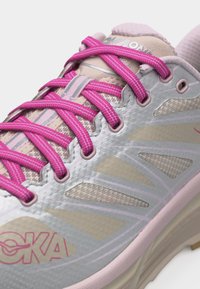 Κοντινό πλάνο αθλητικού παπουτσιού σε ανοιχτό γκρι και μπεζ με φωτεινά ροζ κορδόνια και λογότυπο, που δείχνει υφή από διχτυωτό ύφασμα και συνθετικά επικαλύμματα.