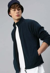 Giacca in pile blu navy con zip completa, colletto alto e tasche laterali; indossata sopra una t-shirt bianca. Texture opaca, design casual.