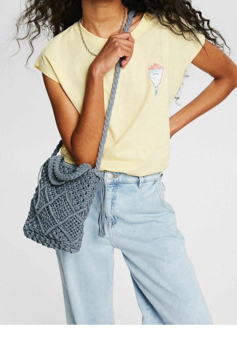 Frau trägt ein gelbes T-Shirt mit Blumenstraußdruck, hellblaue Jeans und hält eine graue, gehäkelte Umhängetasche mit geflochtenem Träger.