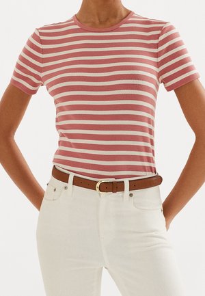 Persoon met een roze-wit gestreept shirt met korte mouwen, een witte jeans en een bruine riem met de handen in de zakken, alleen de romp is zichtbaar.