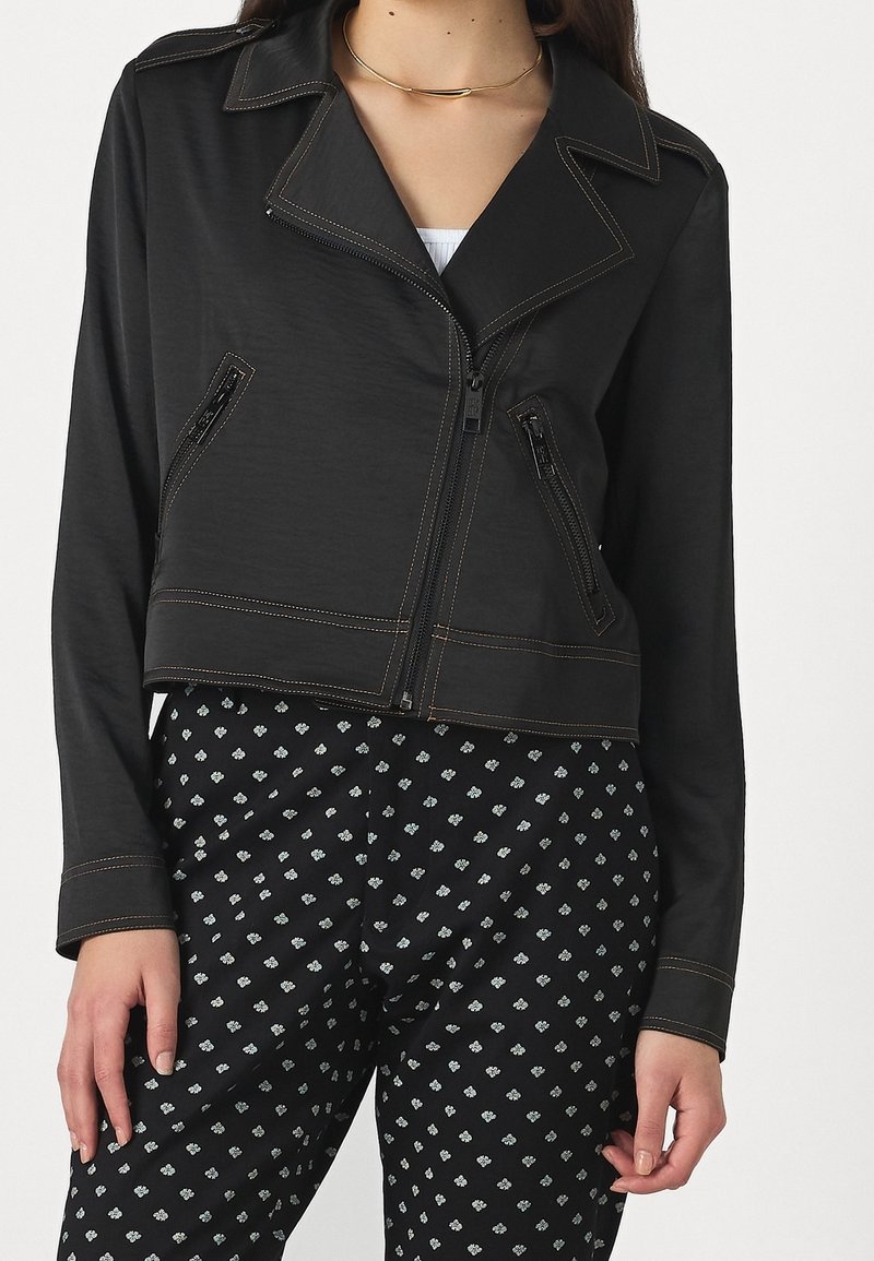 Femme portant une veste courte noire avec des coutures contrastantes et un pantalon noir avec un petit motif floral blanc.