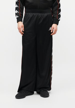 Isik, kes kannab lahtisi musti dressipükse valgete logodega külgedel ja mustasid tenniseid, käed mõlemal pool rahulikult rippumas.