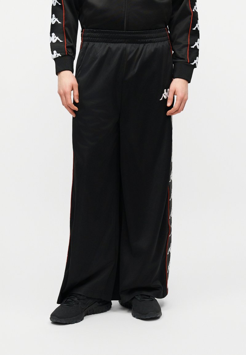 Άτομο που φοράει φαρδιά μαύρη φόρμα με λευκά σχέδια λογότυπου στις πλευρές και μαύρα αθλητικά παπούτσια, με τα χέρια να ξεκουράζονται δίπλα στο σώμα.