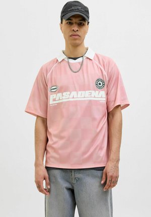 Rosa sports trøje med hvid krave, prydet med et ternet mønster og teksten "PASADENA". Bæret med lysegrå jeans og en kæde halskæde.