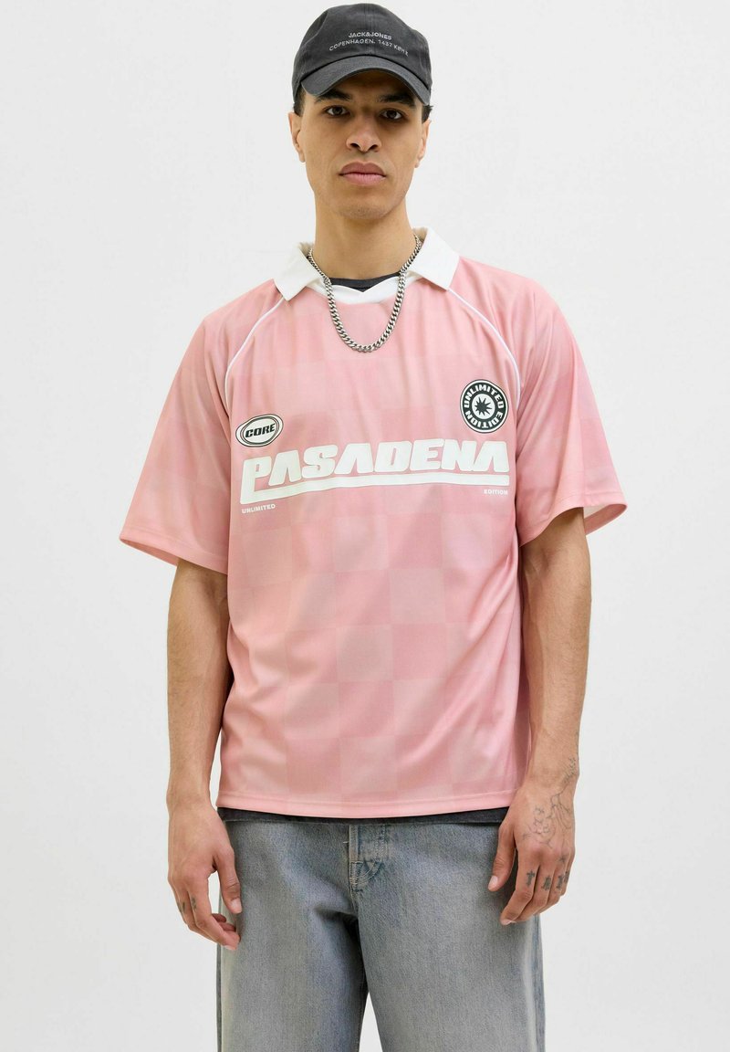 Rosa sports trøje med hvid krave, prydet med et ternet mønster og teksten "PASADENA". Bæret med lysegrå jeans og en kæde halskæde.