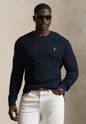 Homme portant un pull torsadé foncé, un pantalon blanc, des lunettes de soleil et une montre-bracelet, debout avec une main dans la poche devant un fond uni.