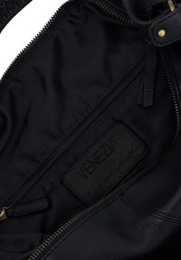 Vnitřek černé kožené kabelky s tmavou textilní podšívkou. Obsahuje zipovou kapsu a koženou značkovou štítku s raženým nápisem "VENEZIA."