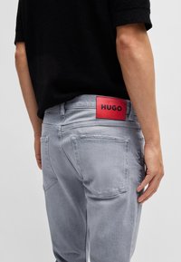 Persona que lleva pantalones vaqueros gris claro con una etiqueta roja de HUGO en la cinturilla y una camiseta negra de manga corta, mostrada desde la parte baja de la espalda hacia abajo.