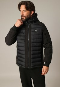 Homme ajustant le col d'une veste matelassée noire à zip avec capuche et logo Deeluxe sur la poitrine, se tenant devant un fond uni.