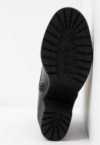 Semelle de bottine noire avec des crampons profonds et stylisés ainsi qu'un petit talon carré, photographiée debout sur une surface blanche.