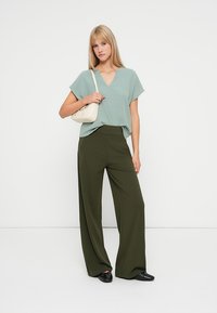 Top verde deschis cu decolteu în v formă și mâneci scurte, asortat cu pantaloni largi verzi măslinii, cu talie înaltă. Modelul poartă o geantă mică crem.