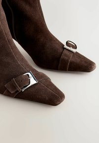 Brązowe zamszowe szpilki z ostro zakończonym noskiem, ozdobione srebrnymi akcentami i detalem z klamrą, charakteryzujące się gładką teksturą i eleganckim wzornictwem.