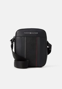 Černá kožená crossbody taška s texturovaným předním dílem, jemnými pruhy v červené a modré barvě a nastavitelným černým popruhem. Na tašce je zobrazeno logo Tommy Hilfiger.