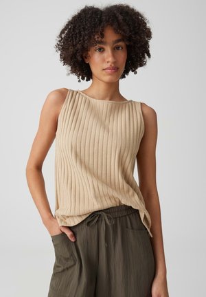Jonge vrouw met krullend haar die een beige mouwloos geribd topje en olijfgroene broek met trekkoord draagt, poseert met hand in zak.