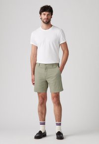 Pantaloni scurți de un verde măsliniu deschis, cu o croială dreaptă, asociați cu un tricou alb cu guler rotund, mocasini negri și șosete în dungi.