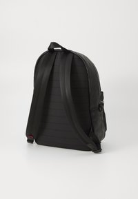 Zaino nero con spallacci regolabili, materiale strutturato e una tasca con zip. Presenta una forma curva e dettagli di pannelli orizzontali.