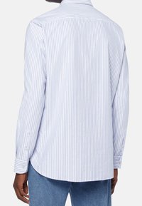 Osoba oblečená v modro-bílém pruhovaném tričku je zachycena zezadu, s dlouhými rukávy a neformálním stylem. Viditelné jsou také džíny.