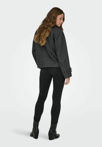 Šedá zkrácená bunda vyrobená z texturované látky, spárovaná s černými skinny džíny a kotníkovými botami. Viděno ze zadní strany, což zdůrazňuje detaily rukávů.