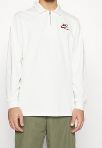 Camisa polo de manga larga blanca con cremallera frontal, que presenta un logo de Nike rojo y negro en el pecho y un diseño de cuello clásico.