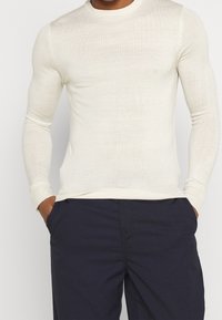 Ljusgräddig långärmad stickad tröja med rund hals och ribbade muddar, med en slät textur och figurnära silhuett. Matchad med marinblå byxor.