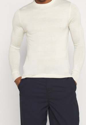 Chemise en tricot à manches longues de couleur crème clair avec un col rond et des poignets côtelés, présentant une texture lisse et une silhouette ajustée. Associée avec un pantalon bleu marine.