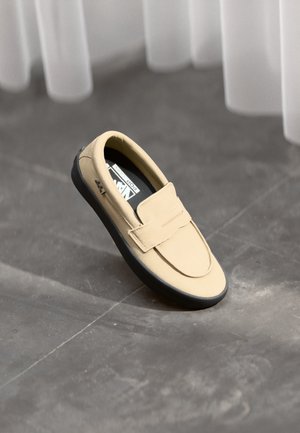 Chaussure beige à enfiler en matériau lisse, dotée d'une large sangle en velcro, d'un bout arrondi, d'une semelle extérieure noire et d'un design minimaliste.