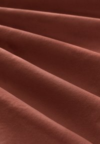 Texturovaná látka v bohaté terakotové barvě, s jemnými žebrovanými vzory a hladkými, skládajícími se vrstvami, které zdůrazňují její jemný materiál.