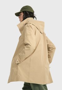 Giacca esterna impermeabile beige con cappuccio, realizzata in materiale liscia. Presenta una vestibilità comoda, tasche laterali e dettagli con marchio sulla manica.
