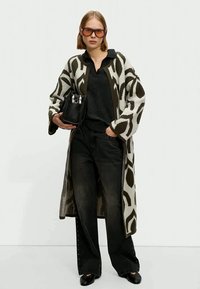 Woman wearing oversized patterned cardigan, black shirt, wide-leg black jeans, black shoes, large sunglasses, holding black handbag.