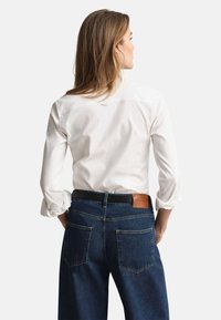 Balti marškiniai ilgomis rankovėmis, rankogaliai sukti, derinami su tamsiai mėlynomis džinsinėmis kelnėmis, kurios turi odinę dėmę ir matomas galines kišenes.