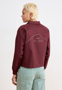 Casaco Nike bordô com gola alta, apresentando um logotipo branco na parte de trás e mangas em contraste. Combinado com calças riscas verdes.