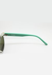 Slnečné okuliare s priehľadnými rámami, tmavými sklami a zelenými plastovými nohami s brandingom "TIKA" a CE značkou na spánku.