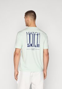 Svetlo metina zelena majica s kratkimi rokavi, ki na hrbtni strani prikazuje drzno mornarsko modro grafiko "MATCH POINT". Gladka tkanina in sproščen kroj.