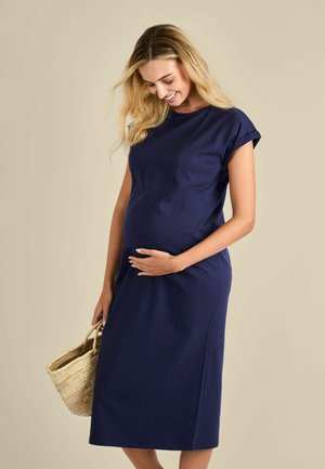 Navykék ruhát viselő terhes nő, aki egy fonott kosarat tart, mosolyogva és lefelé nézve, egy egyszínű bézs háttér előtt áll.