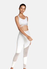 Weißer Sport-BH mit mit Logo verzierten Trägern, kombiniert mit hochtaillierten weißen Leggings, die durchsichtige Einsätze und Farbblock-Akzente aufweisen.
