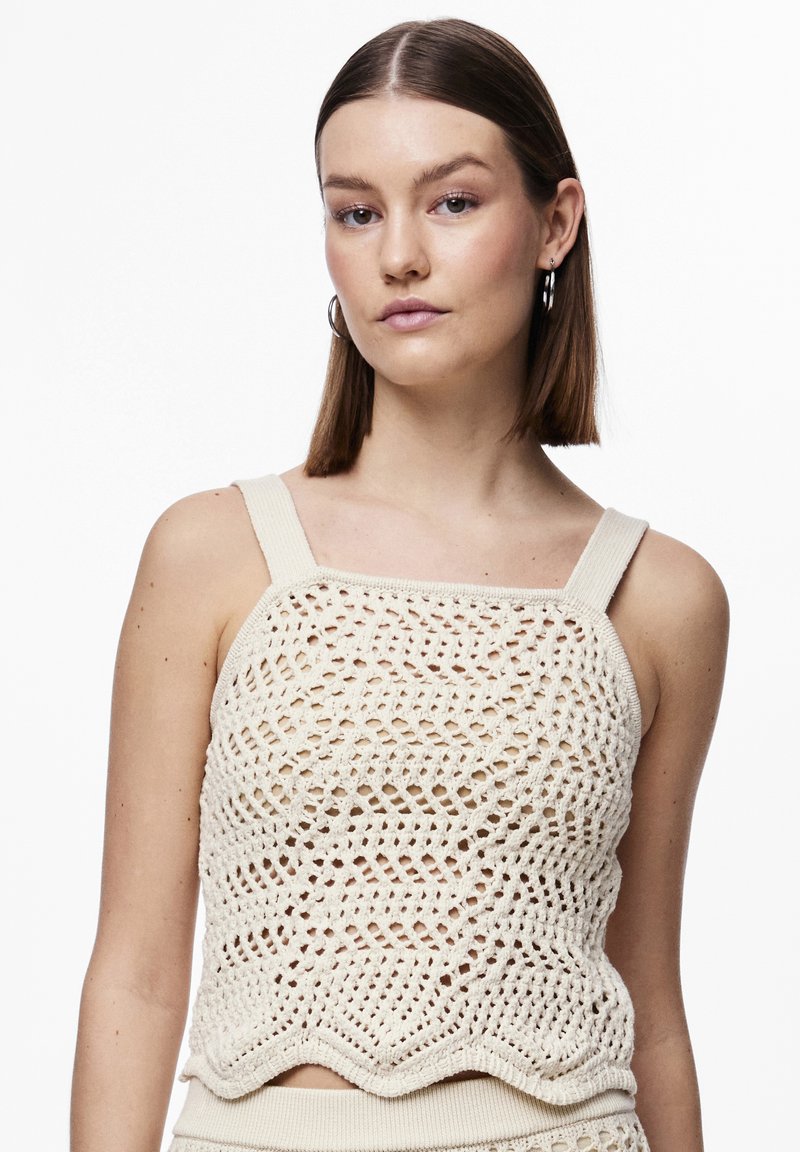 Kremasti pleteni crop top s valovitim rubom, koji sadrži otvorene uzorke i debele, ravne naramenice. Tekstuiran i prozračan materijal.