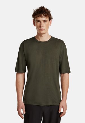 Man met krullend haar, gekleed in een donkergroen T-shirt met korte mouwen en een ronde hals, en een zwarte broek, staand tegen een effen witte achtergrond.