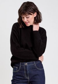 Jeune femme aux cheveux bruns courts portant un pull noir et un jean bleu foncé, regardant vers le bas avec une main près de son cou.