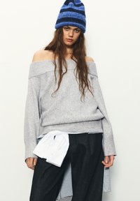 Grauer Schulterfreier Pullover aus weichem Strickmaterial, kombiniert mit dunkler Jeanshose und einem weißem Hemd, das um die Taille gebunden ist. Blau gestreifte Mütze.