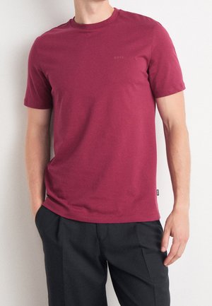 Homme portant un t-shirt à manches courtes uni bordeaux et un pantalon noir, debout avec une main dans la poche contre un mur blanc.