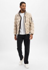 Doudoune matelassée beige avec un col haut, des zips noirs et des poches latérales, portée sur un t-shirt blanc et associée à un jean noir.