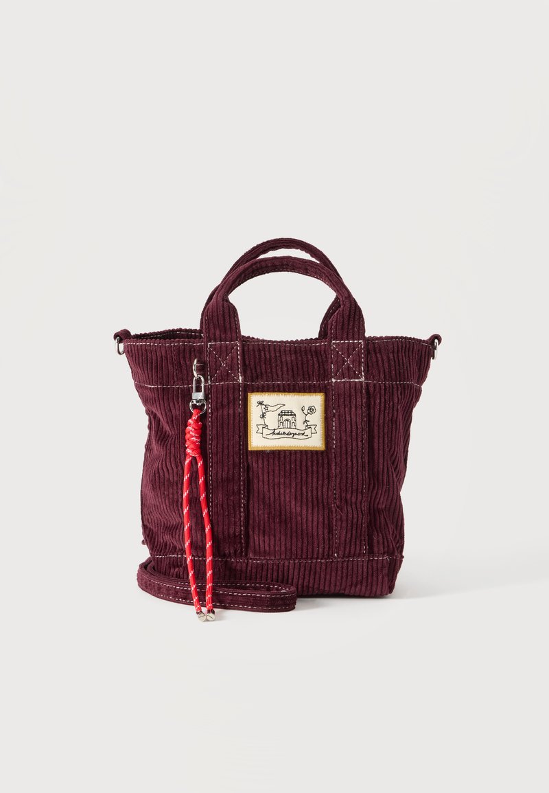 Petit sac cabas en velours côtelé bordeaux avec double poignée, étiquette patch à l'avant et bandoulière tressée rouge attachée à un clip métallique.