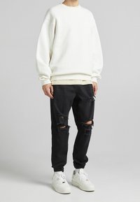 Person med vit sweatshirt, lager på lager beige skjorta, rivna svarta jeans och vita sneakers, som står framför en enkel grå bakgrund.