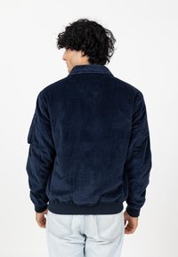 Marineblauwe corduroy bomberjack met een kraag, ribgebreide manchetten en zoom; bevat verticale textuur en een relaxte pasvorm. Achteraanzicht weergegeven.