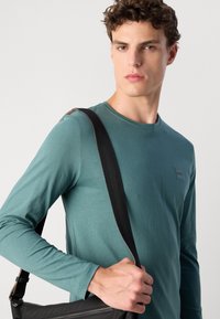 Maglietta a maniche lunghe color teal con collo rotondo e un piccolo logo; il modello tiene in mano una borsa nera con una tracolla testurizzata.
