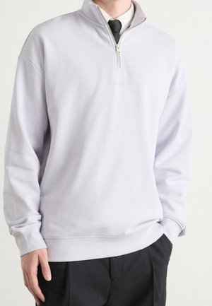 Mann trägt hellgrauen Sweatshirt mit Viertelreißverschluss über weißem Hemd und schwarzer Krawatte, dazu schwarze Hose, steht mit entspannten Händen.