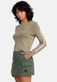 Olijfgroene blouse met lange mouwen, versierd met witte bloemenborduursels en een gouden logo, gecombineerd met een hoge groene cargorok met een opgestikte zak.