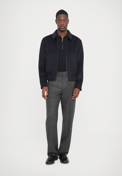 Marineblå zip-up jakke med stor krave; båret over en sort top. Grå mønstrede bukser og sorte sko fuldender looket. Minimalistisk design.