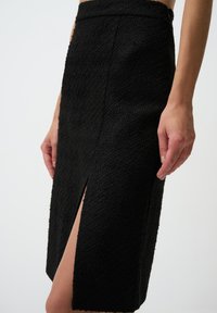 Jupe crayon noire, texturée, avec une fente haute sur la cuisse et une taille ajustée, mettant en valeur un ourlet brut et un design minimaliste.