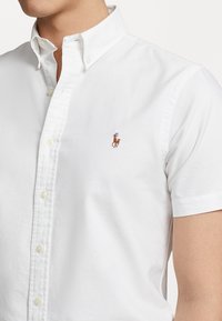 Man i en vit kortärmad skjorta med knappar och en liten brun och blå polospelare-logotyp på bröstet.